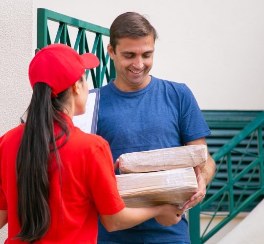 Smiling male customer receiving parcels. Long-haired deliverywoman giving cardboard boxes to happy customer. Female courier in red shirt delivering order and talking. Delivery service and post concept